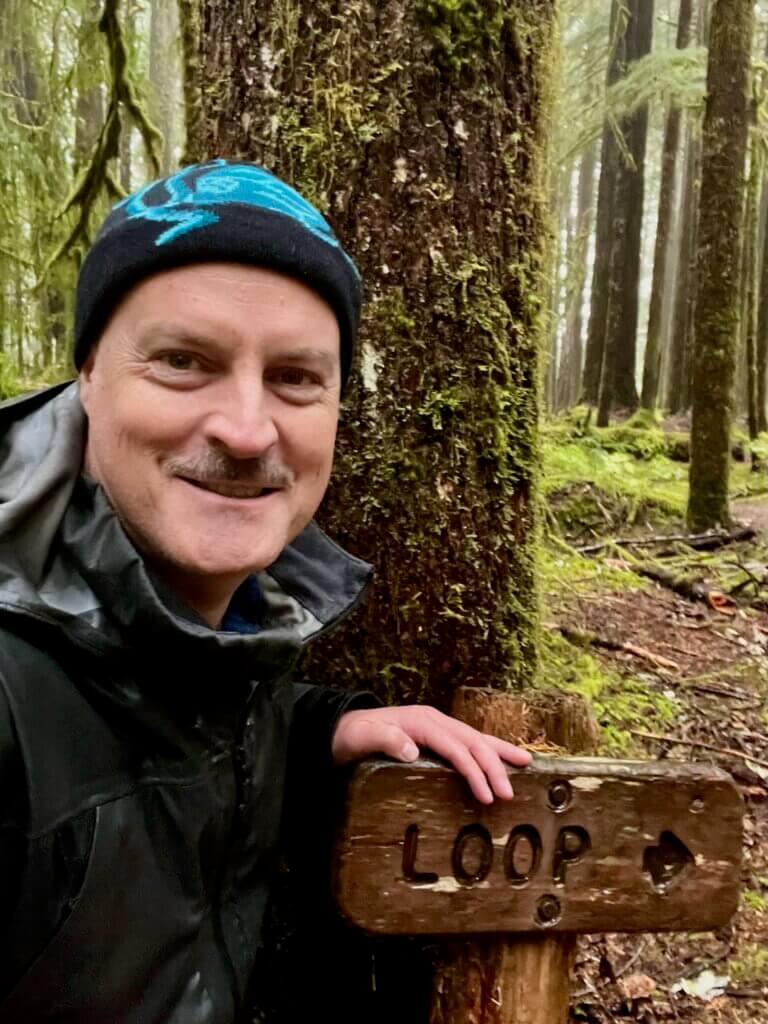 Matthew Kessi gets ready to begin leading people on a forest bathing experience in a wet environment. He's leaning on a sign that says "loop" with an arrow pointing the way to the trial. He's smiling and wearing a black beanie with a blue octopus and a drenched raincoat.