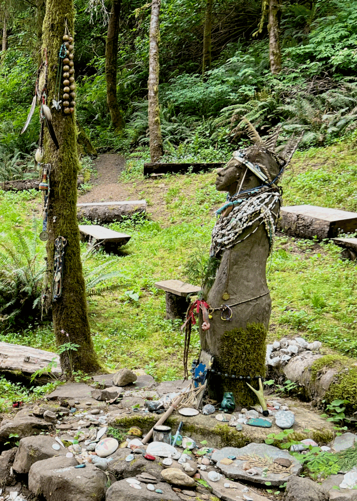 A statue pays tribute to the memory of Amanda at a grotto near Yachats on the Oregon Coast. She is adorned with necklaces made from shells and stones and more trinkets are laid at the base of the statue, which is on stones. There is a lot of greenery and forest behind her.
