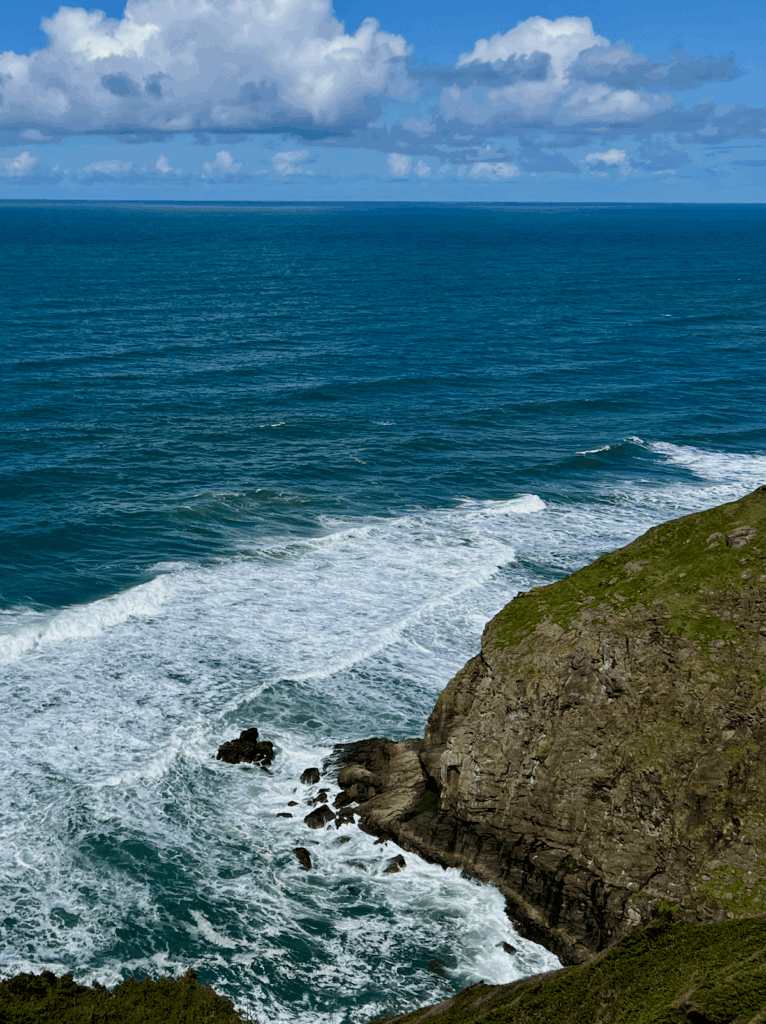 A view of the Pacific Ocean from Highway 101 on the Oregon Coast, near Cape Perpetua. The white foam of the surf is crashing against black rocks as puffy clouds float through a blue sky.