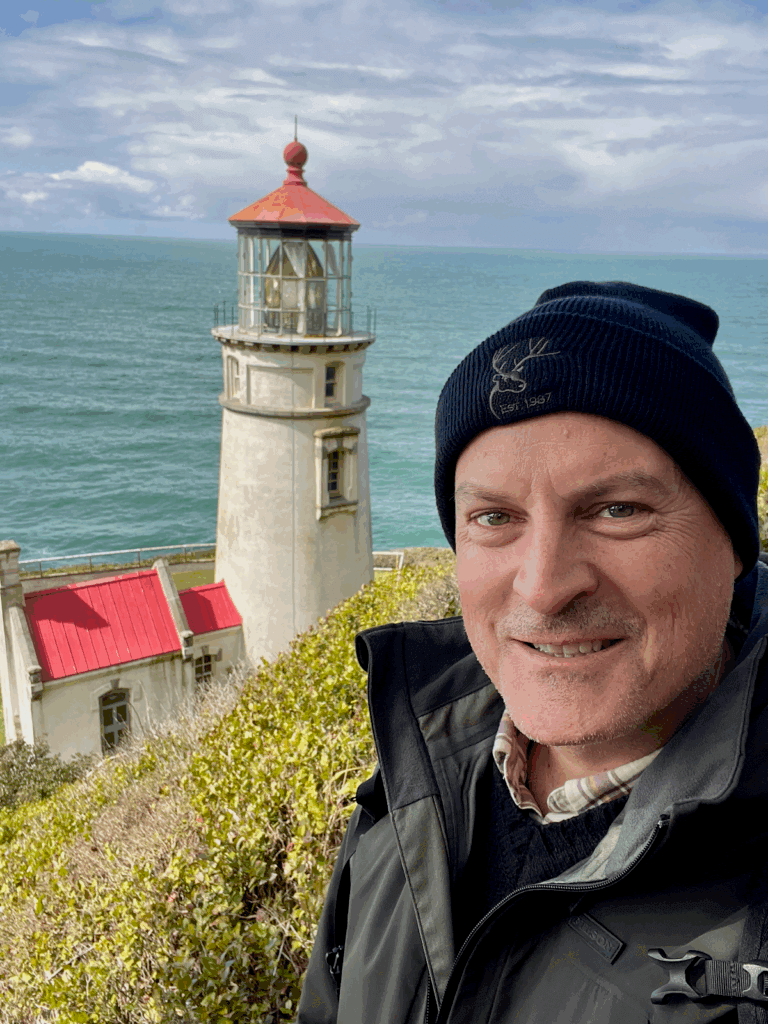 Matthew Kessi poses for a selfie above Heceta Head Lighthouse. He's smiling and wearing a black beanie and dark green jacket. The lighthouse has a red roof and white concrete siding and looks out over blue-green seawater under cloudy skies.
