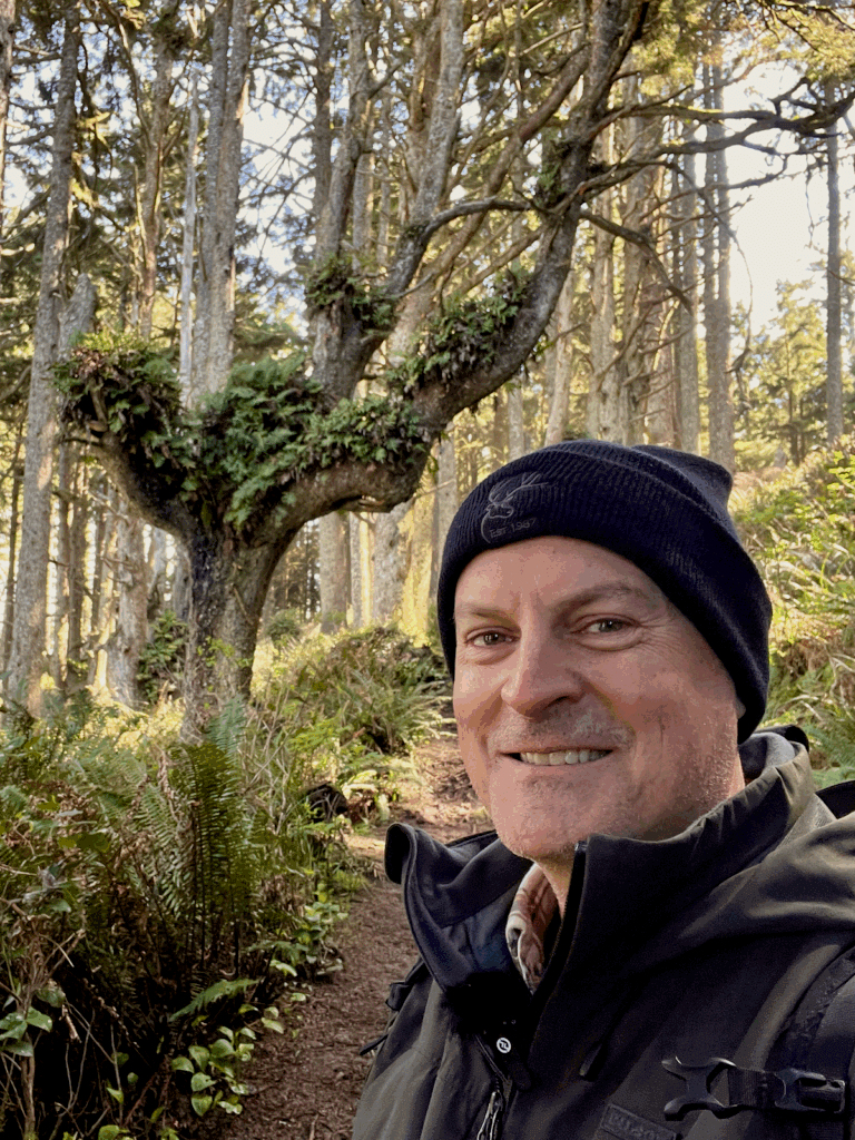 Matthew Kessi poses for a selfie on the Hobbit Trail along Highway 101 on the Oregon Coast. He's smiling and wearing a black beanie and dark jacket. Behind him are wild trees with ferns growing in the branches and various green textures of salal, ferns, and grasses.