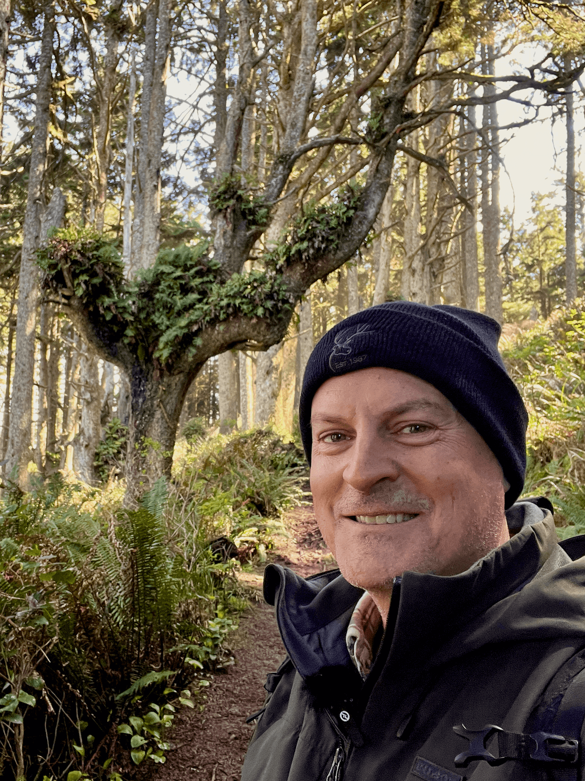 Matthew Kessi poses for a selfie on the Hobbit Trail along Highway 101 on the Oregon Coast. He's smiling and wearing a black beanie and dark jacket. Behind him are wild trees with ferns growing in the branches and various green textures of salal, ferns, and grasses.