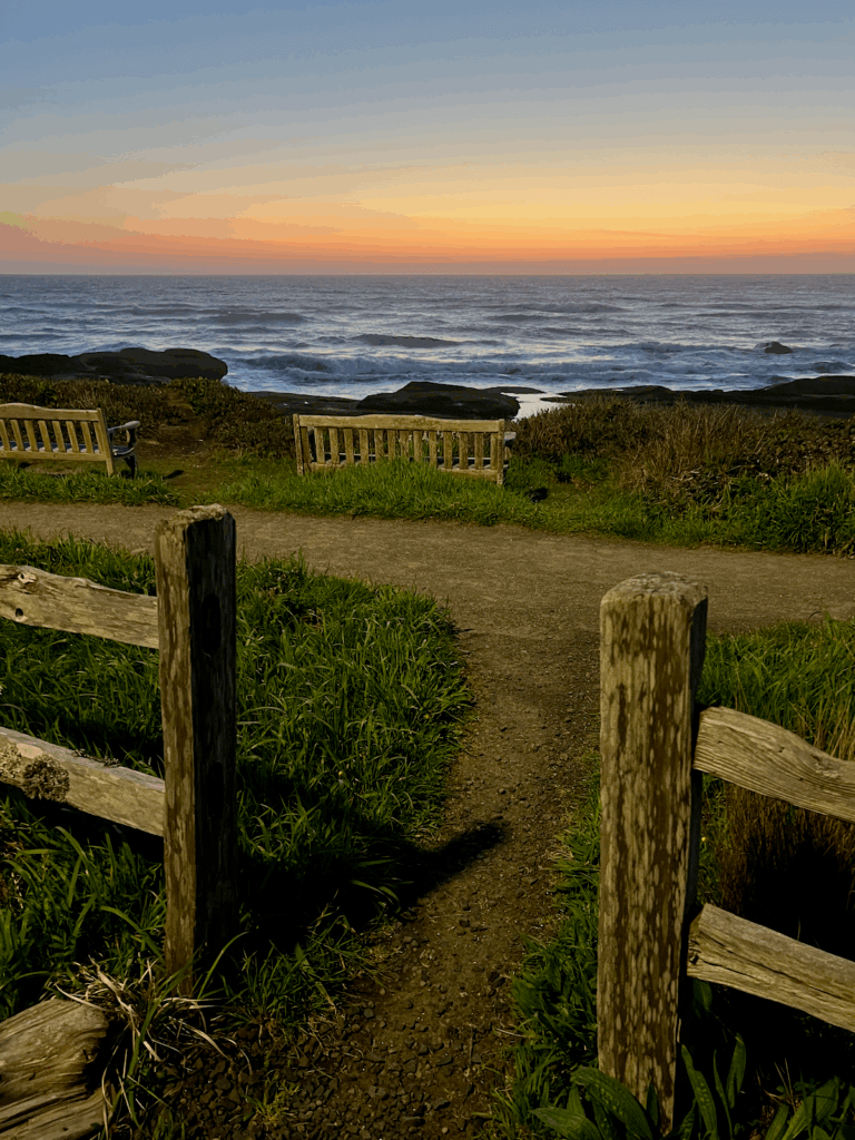 View from a room at the Overleaf Lodge in Yachats Oregon shows peach and pinks of the sunset over the Pacific Ocean. There are two benches in the distance overlooking waves crashing on the rocks nearby.