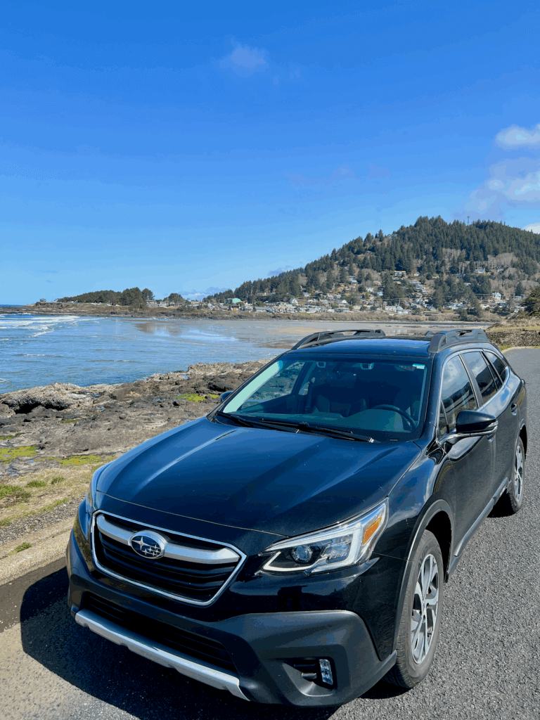 A subaru is pulled up to the edge of a rocky cliff overlooking the beach at Yachats Oregon with the town rising up the hill in the background.