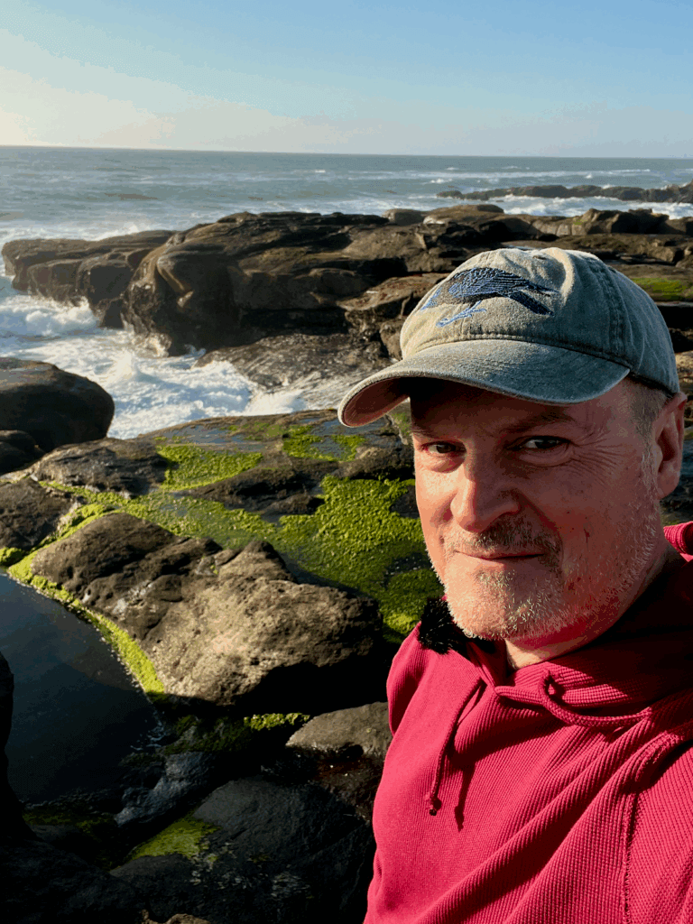 Matthew Kessi poses for a selfie along the 804 Trail in Yachats Oregon. The waves are creeping through breaks in the black rocks while seaweed clings to other rocks.