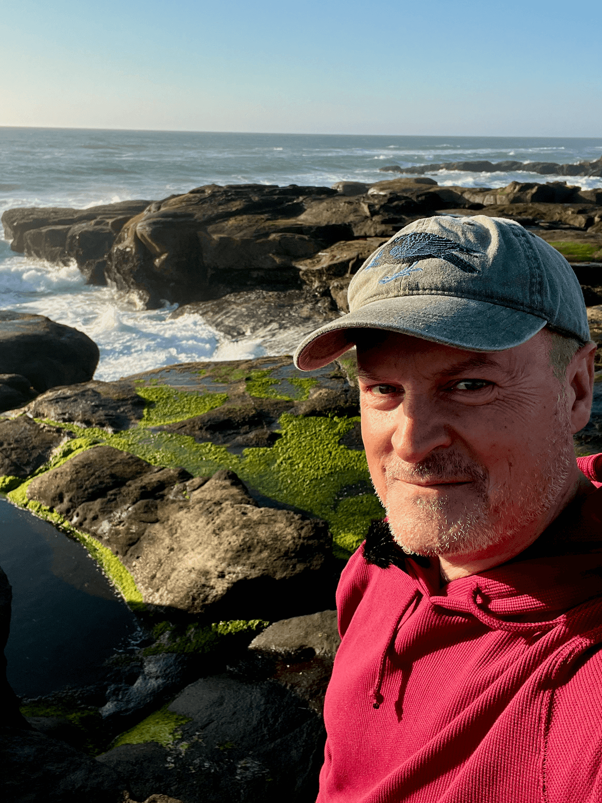 Matthew Kessi poses for a selfie along the 804 Trail in Yachats Oregon. The waves are creeping through breaks in the black rocks while seaweed clings to other rocks.