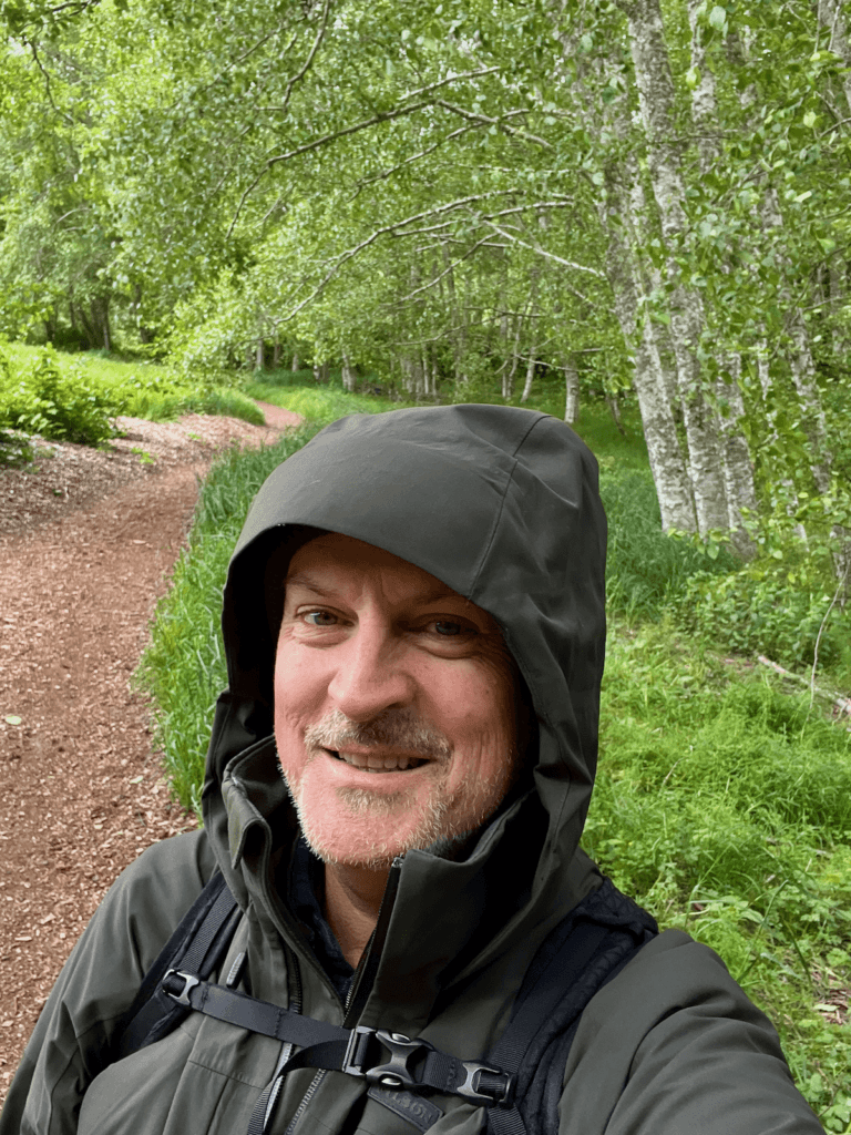Matthew Kessi smiles as he practices the Kinship Questions featured in his book The Wild Cure, on a healing path under newly leafed birch trees on a wood mulch path among grasses. He's wearing a dark green raincoat and a backpack straped to his chest.