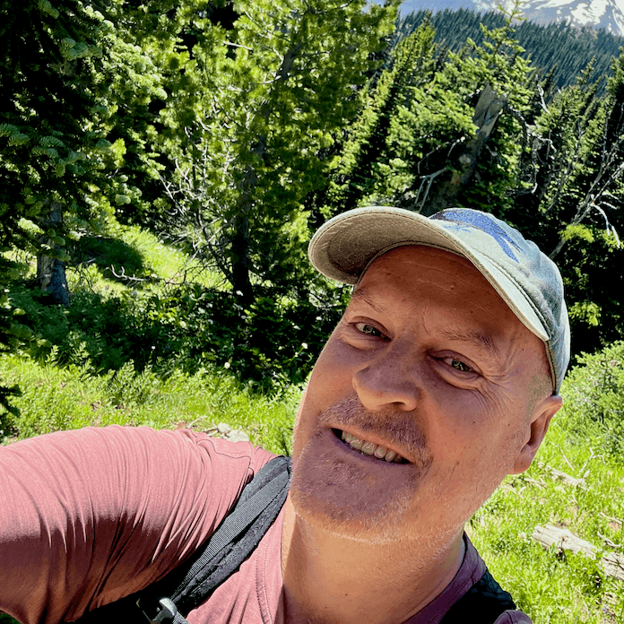 Matthew kessi smiles as he is hiking on Mt. Rainier near Seattle. He's wearing a purple shirt an a green hat with a bird on it.